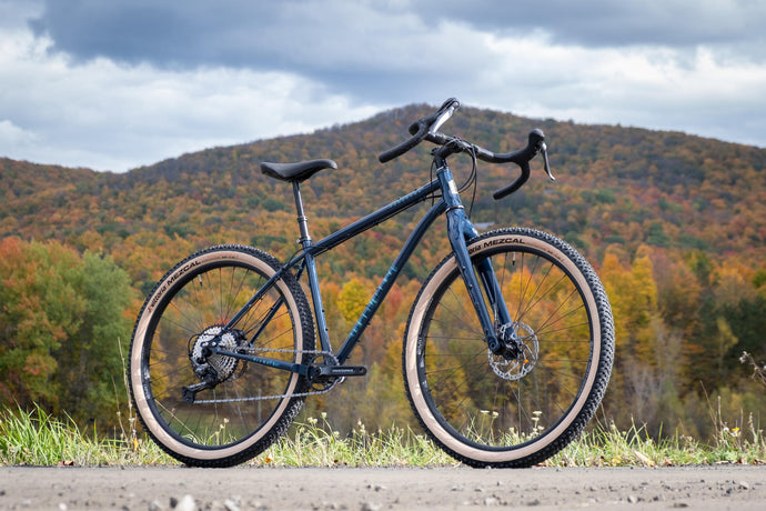 Black touring bike with drop handlebars and knobby tires on a dirt road with fall foliage and hills background