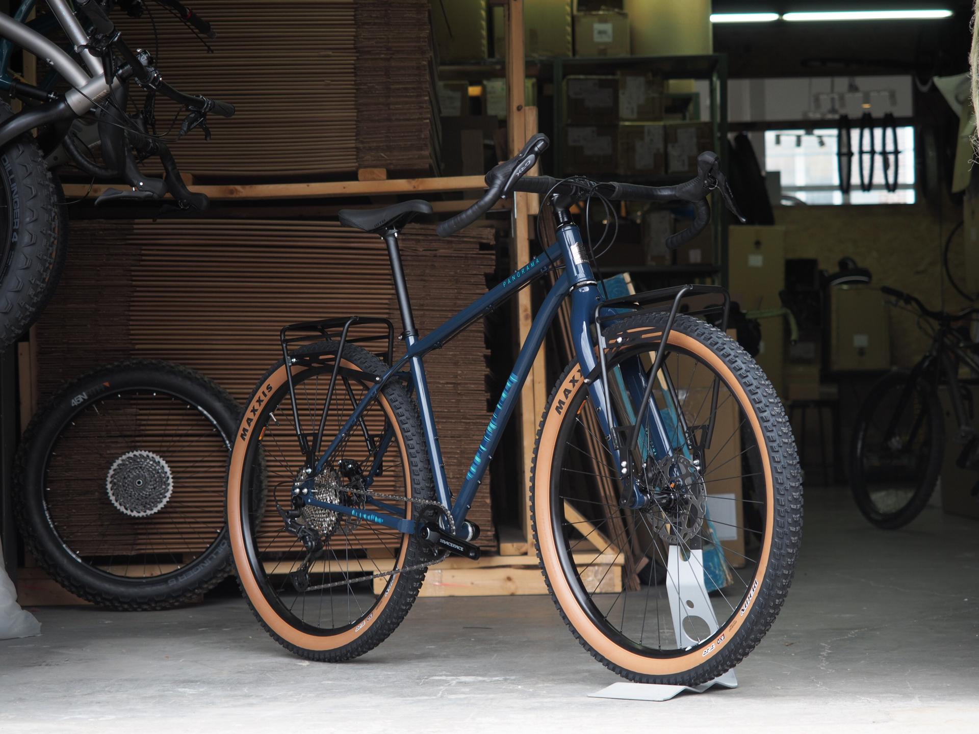 Blue touring bike with tan knobby tires and rear rack standing indoors on concrete floor with stacked cardboard boxes behind
