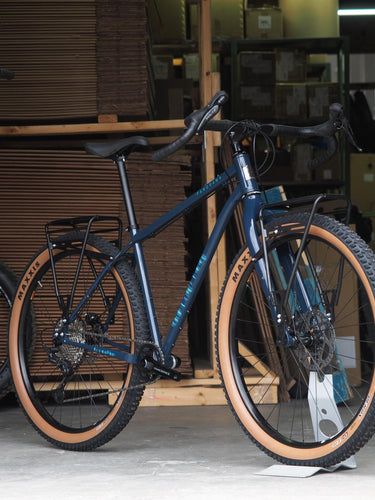 Blue touring bike with tan knobby tires and rear rack standing indoors on concrete floor with stacked cardboard boxes behind