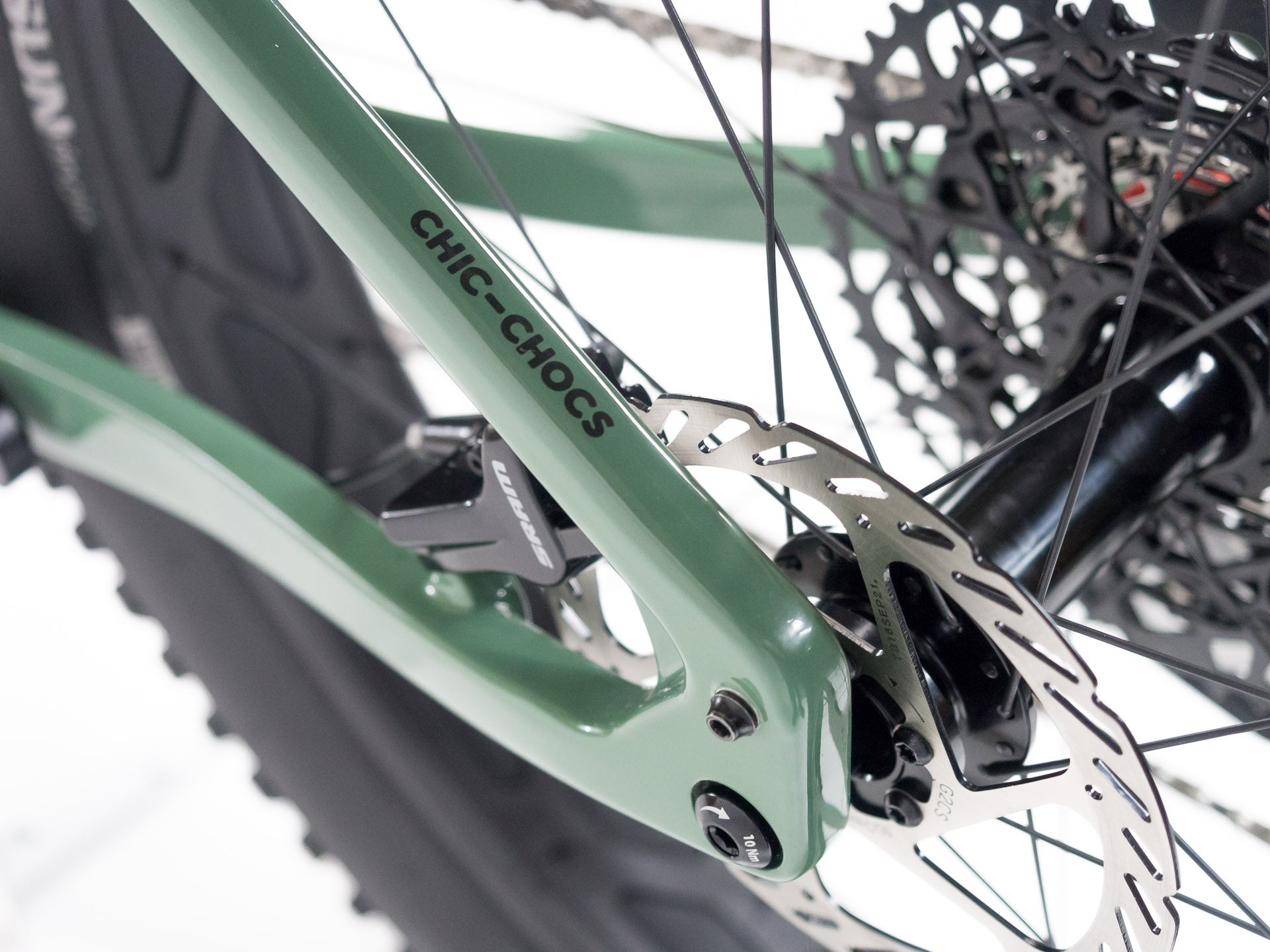 Close-up of green Chic-Chocs bike frame showing rear disc brake, cassette, and wheel spokes on white background